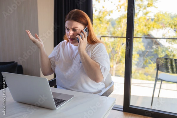 Fototapeta Angry woman sitting at desk with laptop, holding smartphone and gesturing in frustration. Concept of bad internet connection, online problems, stressful work, digital communication issues, remote job
