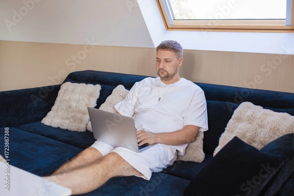 Fototapeta Young man sitting on sofa with laptop and smartphone, working remotely or managing online tasks. Concept of freelancing, digital lifestyle, mobile technology, e-banking, online shopping, communication