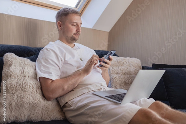 Fototapeta Young man sitting on sofa with laptop and smartphone, working remotely or managing online tasks. Concept of freelancing, digital lifestyle, mobile technology, e-banking, online shopping, communication