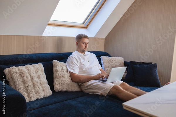 Fototapeta Young man sitting on sofa with laptop and smartphone, working remotely or managing online tasks. Concept of freelancing, digital lifestyle, mobile technology, e-banking, online shopping, communication