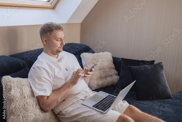 Fototapeta Young man sitting on sofa with laptop and smartphone, working remotely or managing online tasks. Concept of freelancing, digital lifestyle, mobile technology, e-banking, online shopping, communication