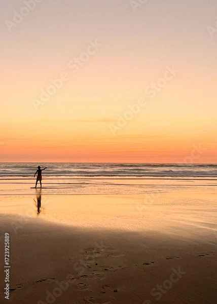 Fototapeta Children play on the sandy shore of Biscarosse as the sun sets, painting the sky in warm hues. The calm waves gently lap at their feet, creating a peaceful atmosphere.