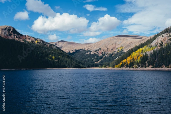 Fototapeta Mountain lake with deep blue water surrounded by forest and autumn foliage under cloudy sky, scenic seasonal landscape