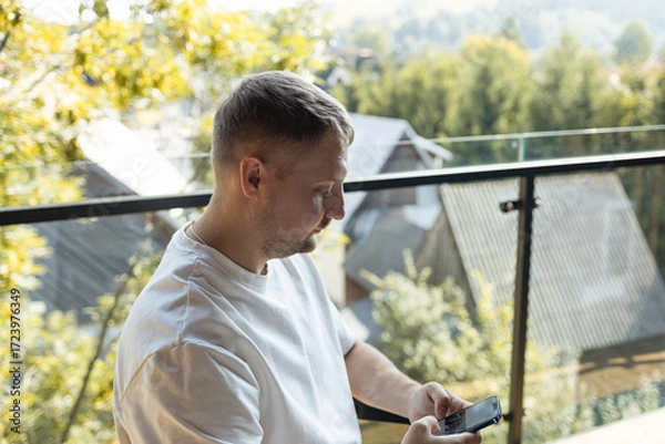 Fototapeta Casual man sitting on balcony in white t-shirt holding smartphone with scenic view of trees and houses in background. Concept of modern lifestyle, technology, remote work, and relaxation.