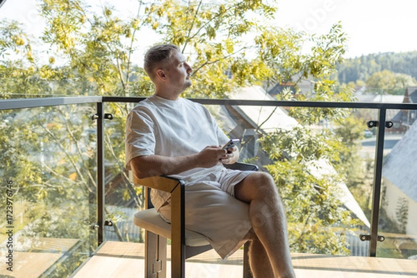 Fototapeta Casual man in white clothes sitting on balcony with coffee cup and smartphone, enjoying peaceful outdoor view of trees and houses. Concept of relaxation, modern lifestyle, and digital connection.