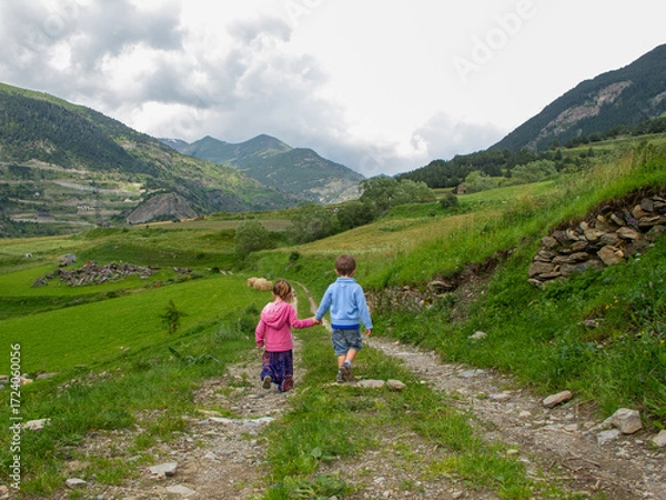 Fototapeta Two children walk hand in hand along a rural path surrounded by green mountains, symbolizing childhood, friendship, and freedom in nature
