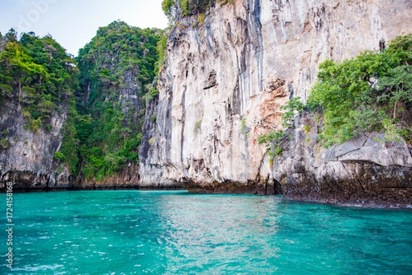 Fototapeta Turquoise water lapping against towering limestone cliffs, adorned with lush vegetation, creates a breathtaking scene in Monkey Bay, Phi Phi Don, Thailand