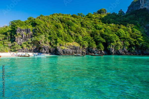 Fototapeta Nui Beach in Krabi, Thailand, featuring a small sandy beach nestled against lush green cliffs with longtail boats floating on clear turquoise water and people enjoying the tropical scenery