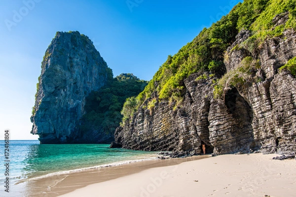 Fototapeta Nui Beach coastline features towering limestone cliffs covered with lush vegetation and a small cave opening onto the sandy beach, with clear turquoise ocean waters