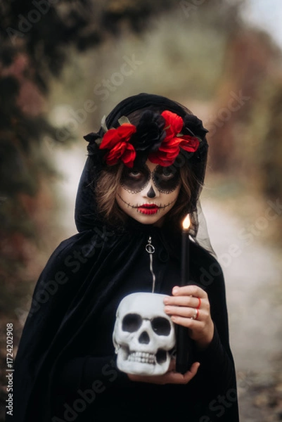 Fototapeta A witch girl with skull makeup holds a candle in a dark forest on Halloween