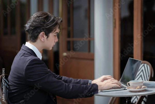 Obraz A young businessman in a black suit works at a coffee shop, using a laptop and a mobile phone while drinking coffee outdoors. The New technology business works.
