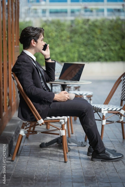 Obraz A young businessman in a black suit works at a coffee shop, using a laptop and a mobile phone while drinking coffee outdoors. The New technology business works.