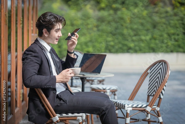 Obraz A young businessman in a black suit works at a coffee shop, using a laptop and a mobile phone while drinking coffee outdoors. The New technology business works.