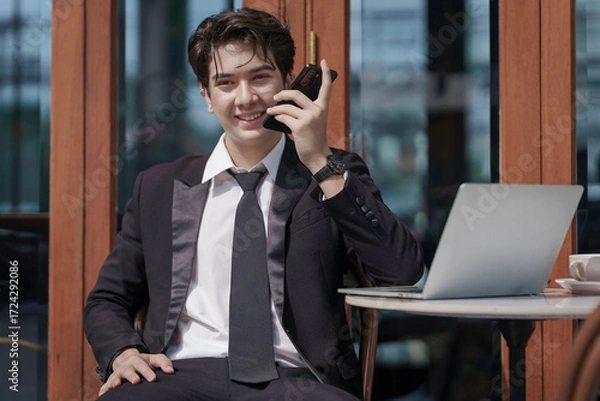 Obraz A young businessman in a black suit works at a coffee shop, using a laptop and a mobile phone while drinking coffee outdoors. The New technology business works.