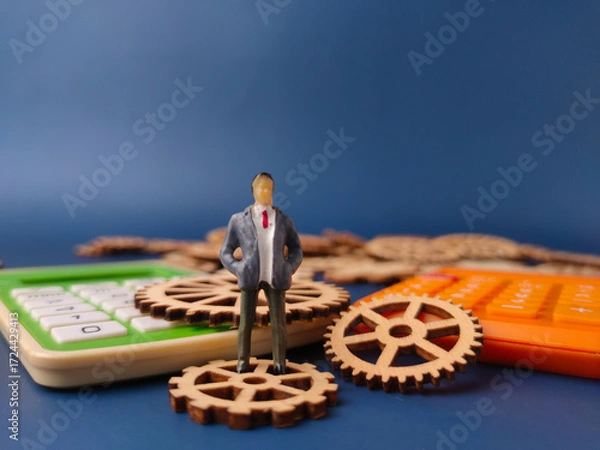 Fototapeta Closeup of a miniature figure of a businessman standing on a wooden gear, with calculators in the foreground