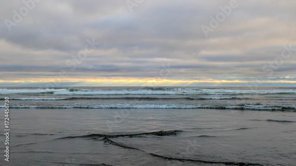 Fototapeta Side view from a wet beach with an emphasis on the small waves rolling in, out westward towards the pacific ocean, under a majestic and complicated cloud-scape near dusk.