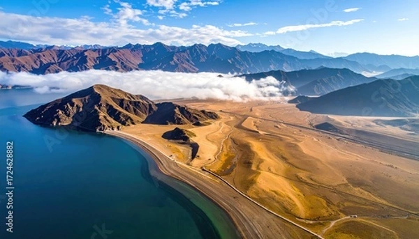 Fototapeta Aerial shot of a desert landscape meeting a calm lake under a sky dotted with clouds, framed by a background of majestic mountains