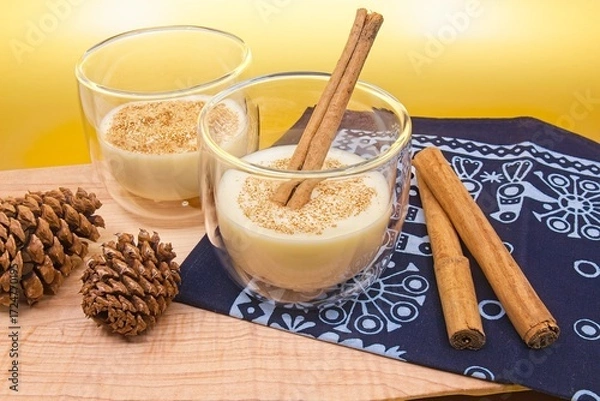 Fototapeta Two cups of eggnog with a cinnamon stick in one glass and cinnamon in both glasses. Pine cones and cinnamon sticks sit on a wood cutting board and blue cloth with holiday decorations. 