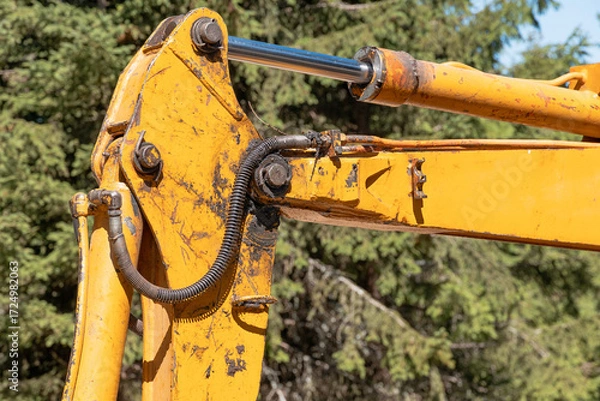 Fototapeta Yellow excavator arm featuring a hydraulic piston and cylinder, showing wear and tear from heavy industrial use in a natural outdoor setting