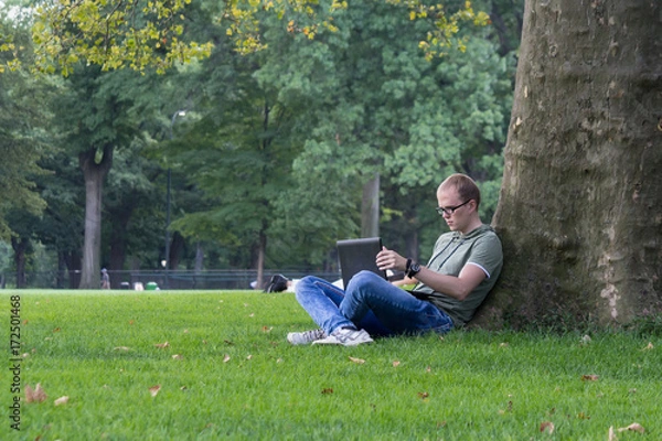 Fototapeta Young man in glasses work on laptop sitting on the grass near the tree in park. Maybe student or hacker