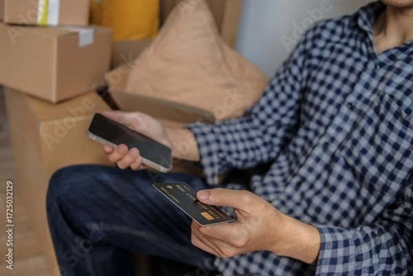 Fototapeta Man Holds Credit Card and Phone Making Payment Amidst Moving Boxes
