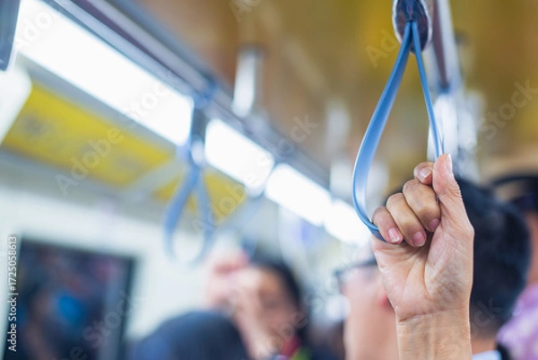 Fototapeta Close up of a person’s hand gripping a hanging strap inside a busy subway train, symbolizing daily commute, transportation, and urban travel.