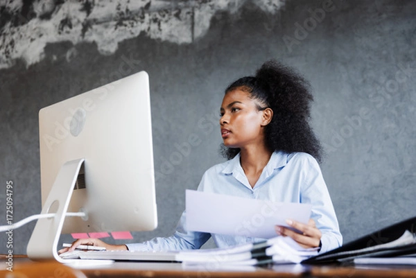 Fototapeta A young professional woman works at her desk with a computer, reviewing charts and opening a file folder, reflecting focus, organization, productivity, and dedication in a modern business environment.