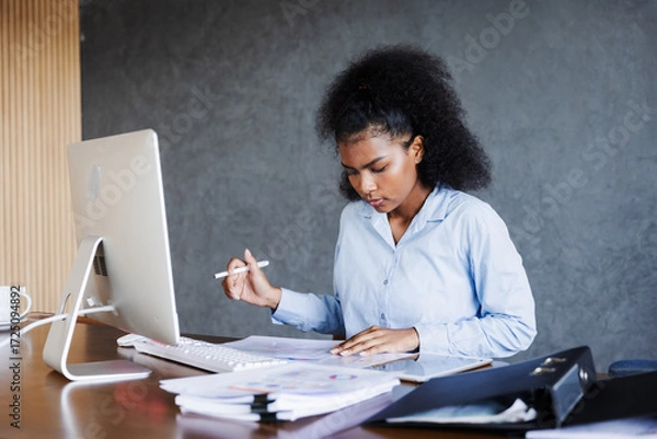 Fototapeta A young professional woman works at her desk with a computer, reviewing charts and opening a file folder, reflecting focus, organization, productivity, and dedication in a modern business environment.
