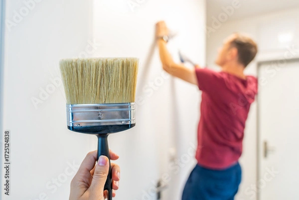 Fototapeta Close-up of a hand holding a paintbrush with a man painting the wall in the background. Concept of manual work, home renovation and attention to detail during interior improvement.