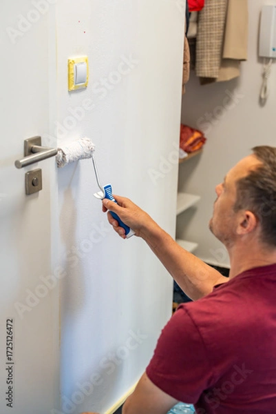 Fototapeta Man painting the wall with a roller in white during home renovation. Interior improvement, surface transformation and DIY work with paint tools.