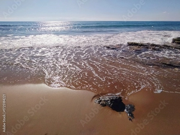 Fototapeta Wet Sand and Foaming Waves on the Beach with Blue Sea and Sun Reflection - Serene Coastal Scene.