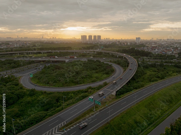 Fototapeta An elevated view of a busy highway interchange in Hanoi at sunset, with cars and trucks navigating the winding ramps and overpasses, all set against a backdrop of a city skyline.
