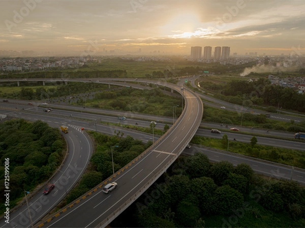 Fototapeta An elevated, wide-angle view of a highway interchange in Hanoi at sunset, showing the sprawling ramps, bridges, and lanes enveloped by lush greenery with distant high-rise buildings on the horizon.