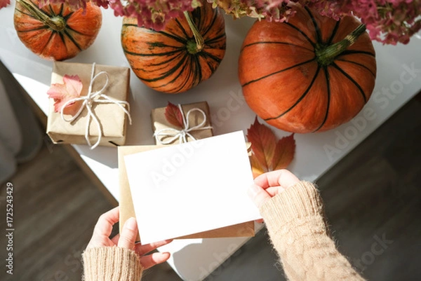 Fototapeta Cozy autumn flat lay with hands in sweater holding blank greeting card, surrounded by pumpkins, wrapped gifts and fall leaves on white surface. Ideal for seasonal greetings and Thanksgiving