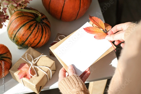 Fototapeta Female hands in cozy sweater hold blank card and autumn leaf, surrounded by pumpkins and gifts on sunlit white table. Perfect for fall designs, invitations and Thanksgiving