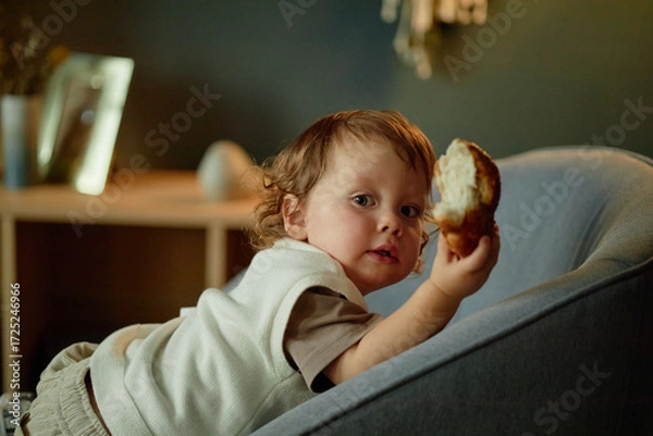 Fototapeta Portrait of Caucasian toddler boy holding piece of bread while sitting on sofa, turning head toward camera