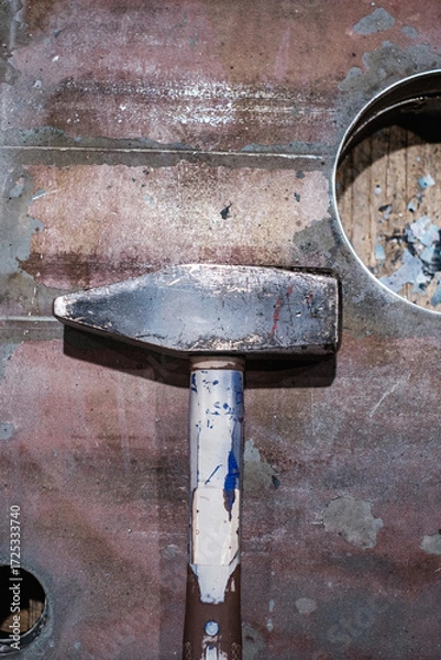 Fototapeta Worn sledgehammer resting on rusty industrial metal surface, close-up of heavy-duty hand tool in workshop with paint marks and cut-out detail