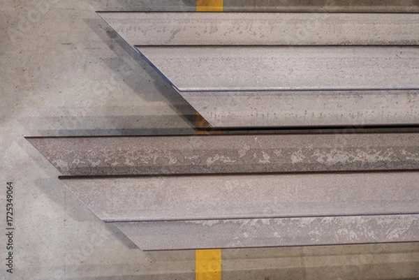 Fototapeta Stacked structural steel profiles in warehouse, close-up of industrial metal beams with worn texture and shadows on concrete floor