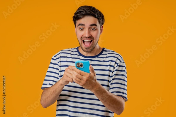 Fototapeta Shocked young Slavic man staring at smartphone on yellow studio with open mouth