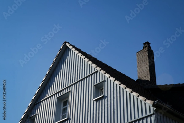Fototapeta Upper part of a blue house with pointed roof, windows and chimney, against a clear blue sky.