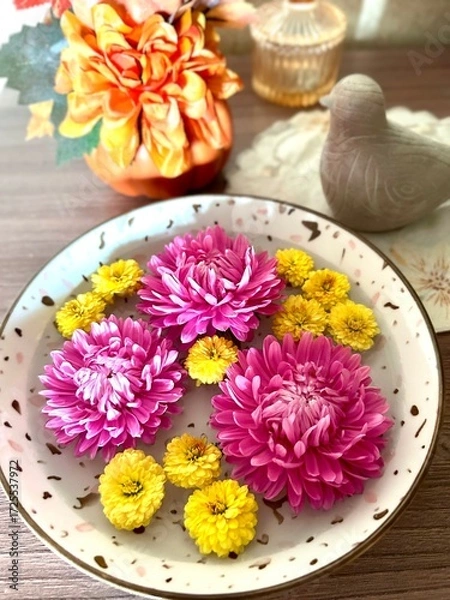 Obraz A serene display of pink and yellow flowers floating in a decorative bowl, complemented by a vibrant orange floral arrangement and rustic ceramic dove on a wooden surface.