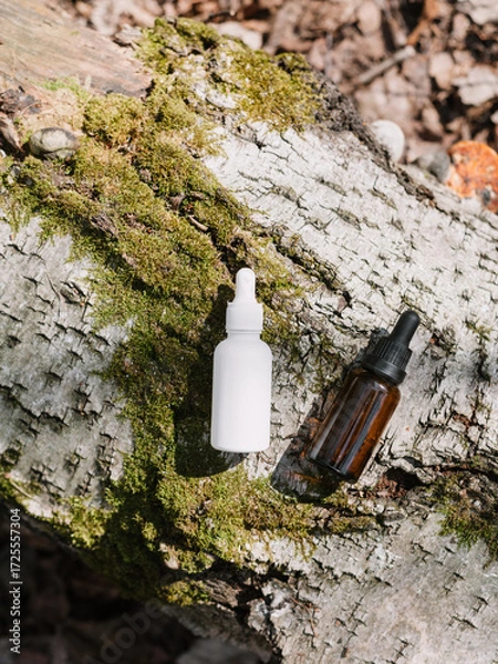 Fototapeta A dark and light glass serum bottle with a dropper, tree bark wood, moss. forest on a sunny day nature background. Concept of skin care, hydration, cosmetic demonstration eco-friendly product mock up