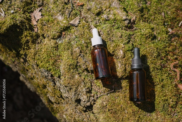 Fototapeta A dark and light glass serum bottle with a dropper, tree bark wood, moss. forest on a sunny day nature background. Concept of skin care, hydration, cosmetic demonstration eco-friendly product mock up