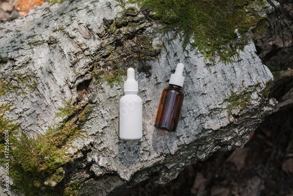 Fototapeta A dark and light glass serum bottle with a dropper, tree bark wood, moss. forest on a sunny day nature background. Concept of skin care, hydration, cosmetic demonstration eco-friendly product mock up