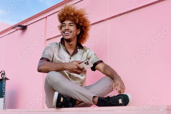 Fototapeta Relaxed African American man with curly ginger hair sitting outdoors holding smartphone, glancing sideways with smile, representing positive attitude toward mobile technology and connectivity.