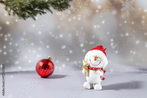 Fototapeta A toy figure of a snowman holding a star and a Christmas tree ball on a festive snowy background. Christmas celebration concept, greeting card