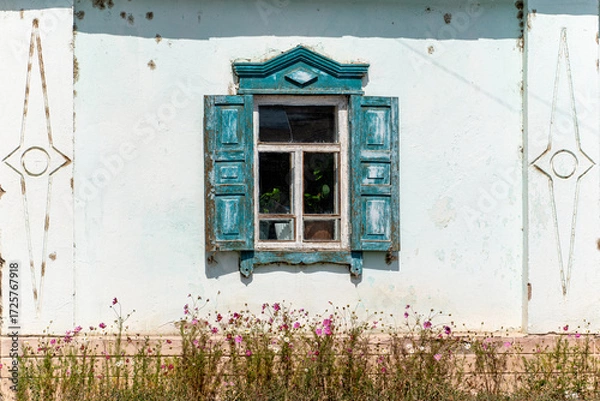 Fototapeta An old, weathered wooden window with peeling blue paint and shutters on a light-colored, textured wall of a traditional house.
