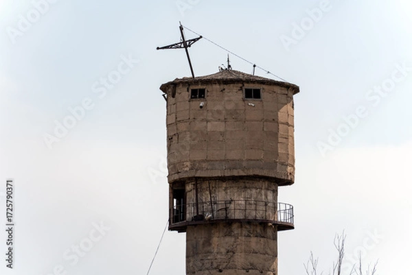 Fototapeta An old, weathered concrete water tower stands against a bright sky, featuring a worn, textured surface and small windows near its top.