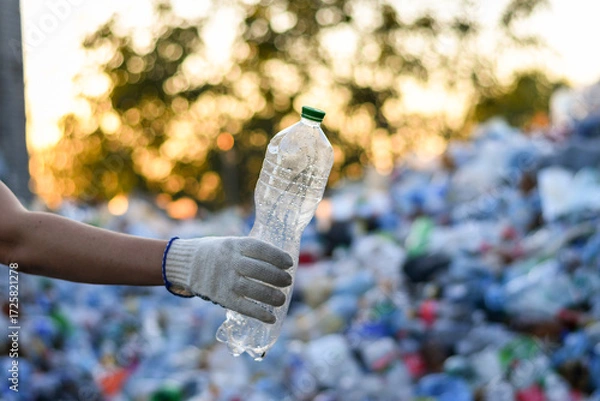 Fototapeta Hand holding empty plastic bottle at recycling center on the blurred background with pile of plastic waste. Selective focus. 