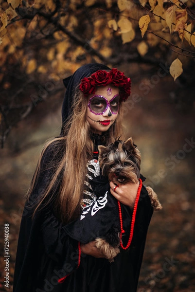 Fototapeta A young woman in a Day of the Dead costume: purple and violet skull makeup, red roses on a wreath, and a black cloak. She held a small dog against the backdrop of an autumn park with fallen leaves.